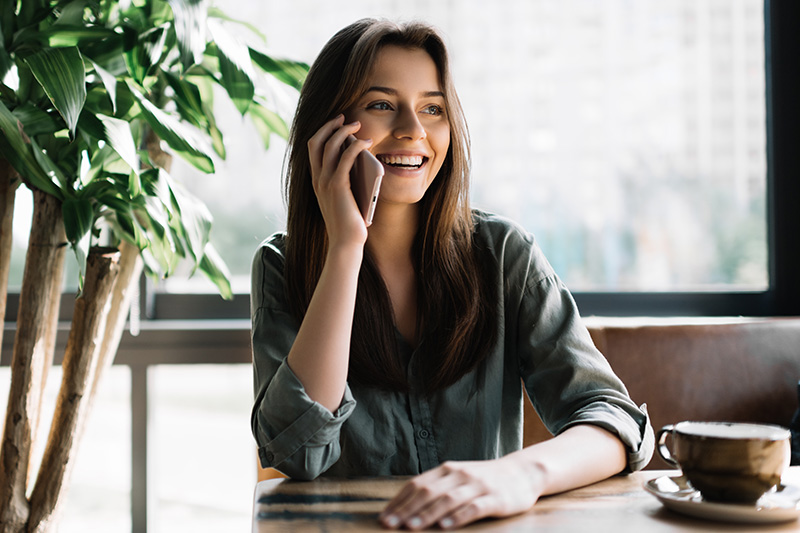 A woman sitting at a table, talking on her phone with a smile, in an indoor setting with potted plants and large windows.